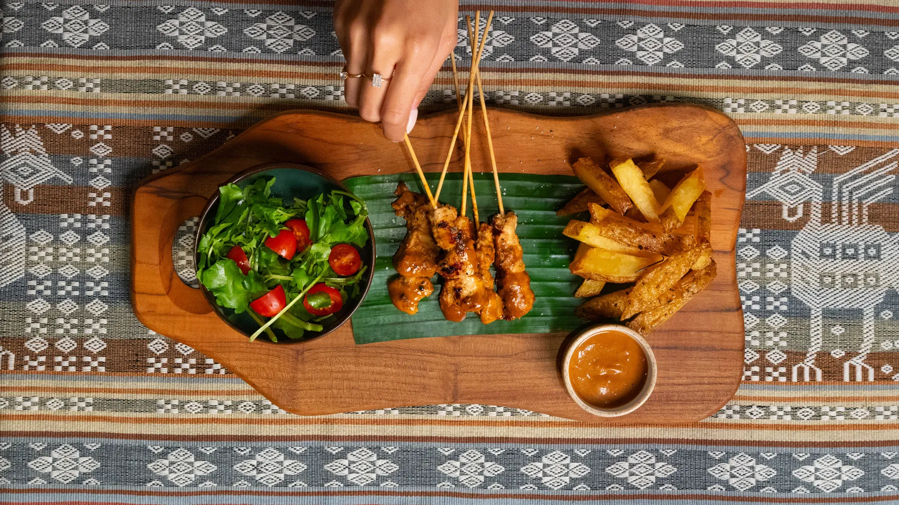Papan sate ayam dengan bumbu kacang, kentang goreng, dan salad di atas alas ikat