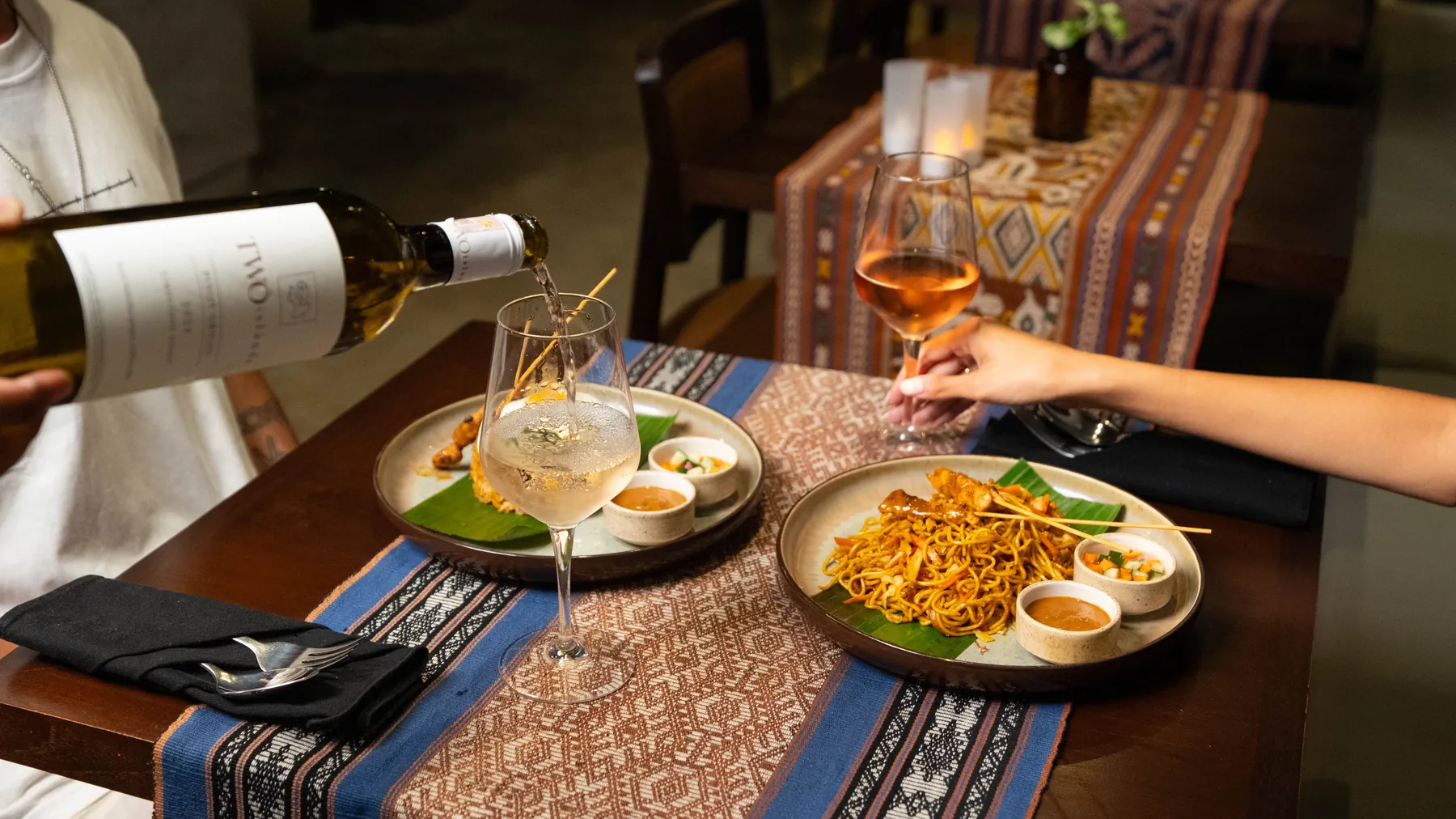 Wine poured at a candlelit table set with mie goreng and satay